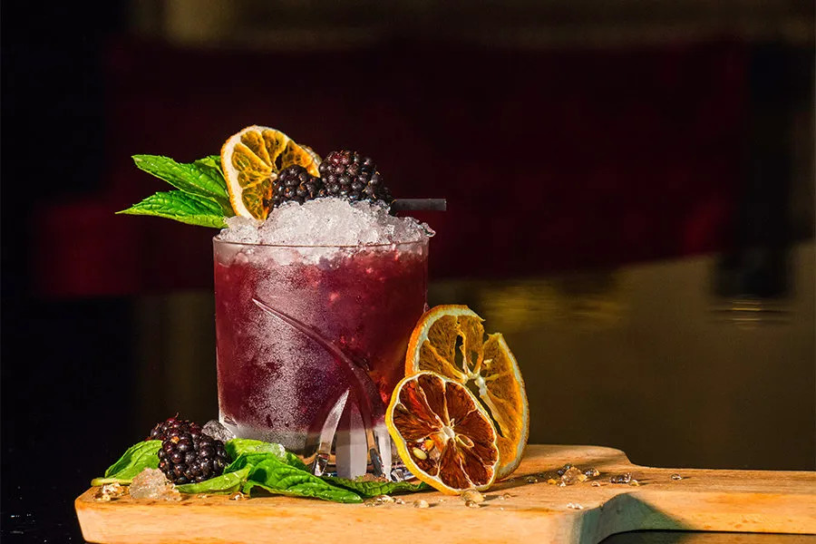 Cóctel morado con moras, hielo picado, rodajas de naranja deshidratada y hojas de menta.