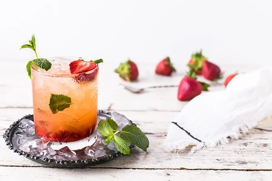 Cóctel con fresas y menta en vaso sobre un plato con hielo, fresas y paño de fondo.
