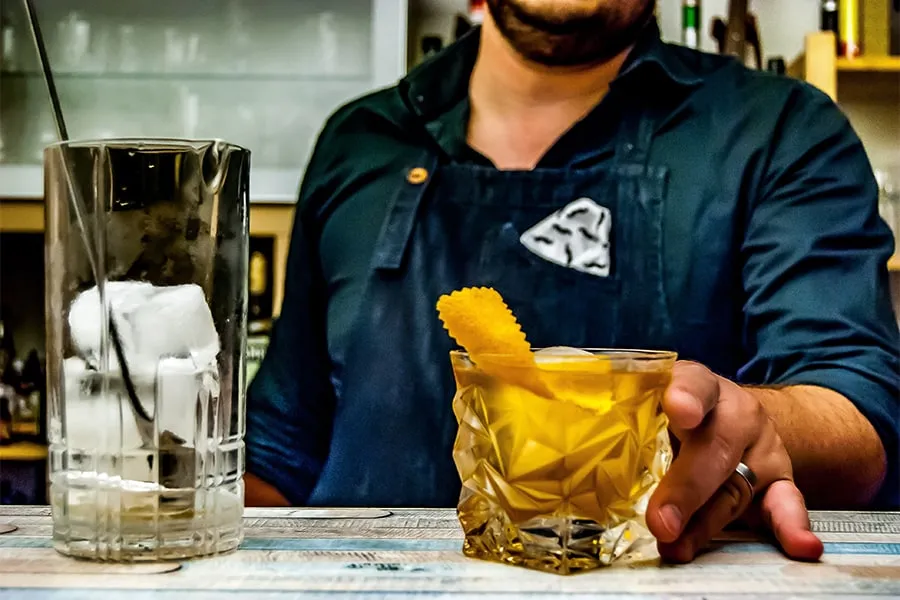 Bartender sirviendo un cóctel en un vaso decorado con cáscara de naranja, junto a un vaso con hielo.