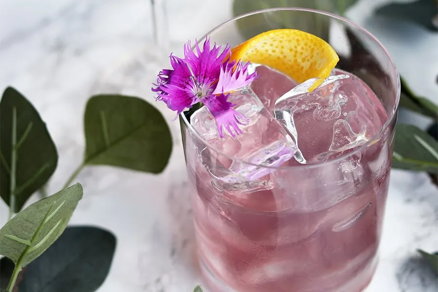 Cóctel rosa con hielo, decorado con una flor morada y rodaja de limón sobre una mesa con hojas verdes.
