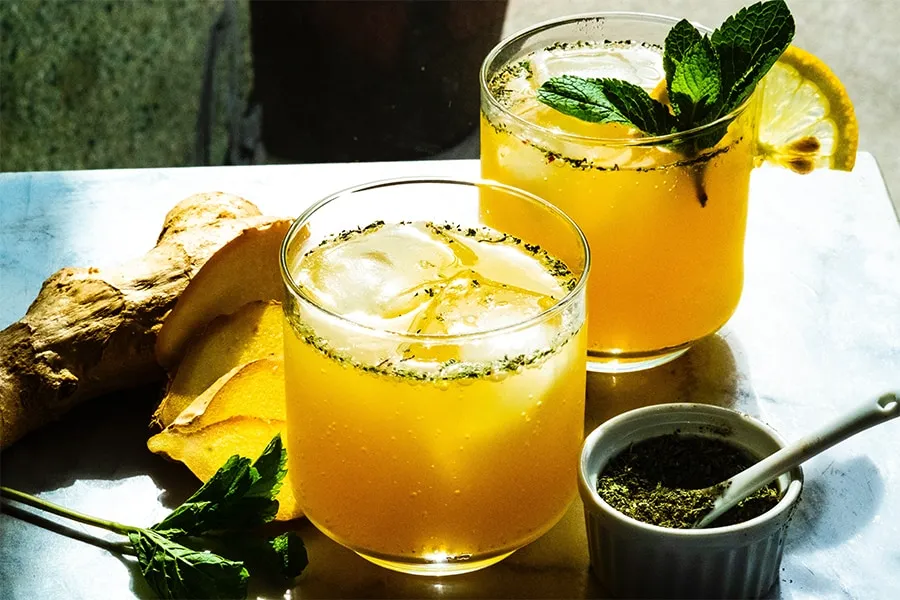 Dos vasos de bebida amarilla con hielo, rodaja de limón y menta, junto a jengibre y hierbas.