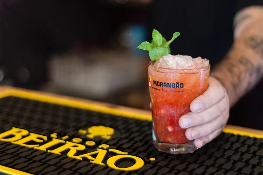 Coctel rojo con menta y hielo en un vaso, sostenido por una mano sobre una barra.