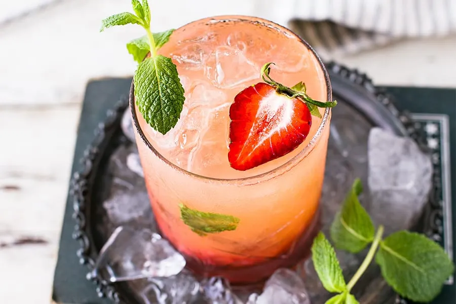 Cóctel rosado con hielo, fresa y menta en bandeja negra.