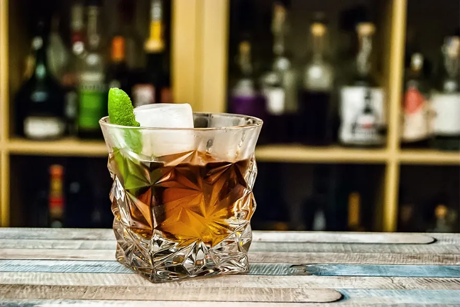 Vaso de cóctel con hielo, rodaja de limón, fondo de botellas borroso.