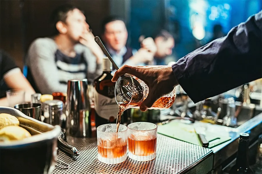 Una mano sirve whisky en dos vasos en un bar, con gente conversando de fondo.