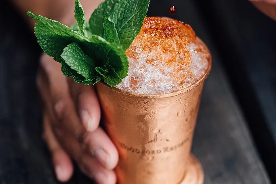 Copa de cobre con cóctel, hielo picado, una gota roja y menta fresca, sostenida por una mano.