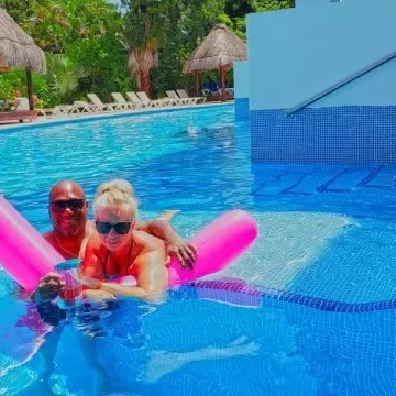 Pareja en piscina con flotador rosa, rodeada de árboles y tumbonas bajo palapas.