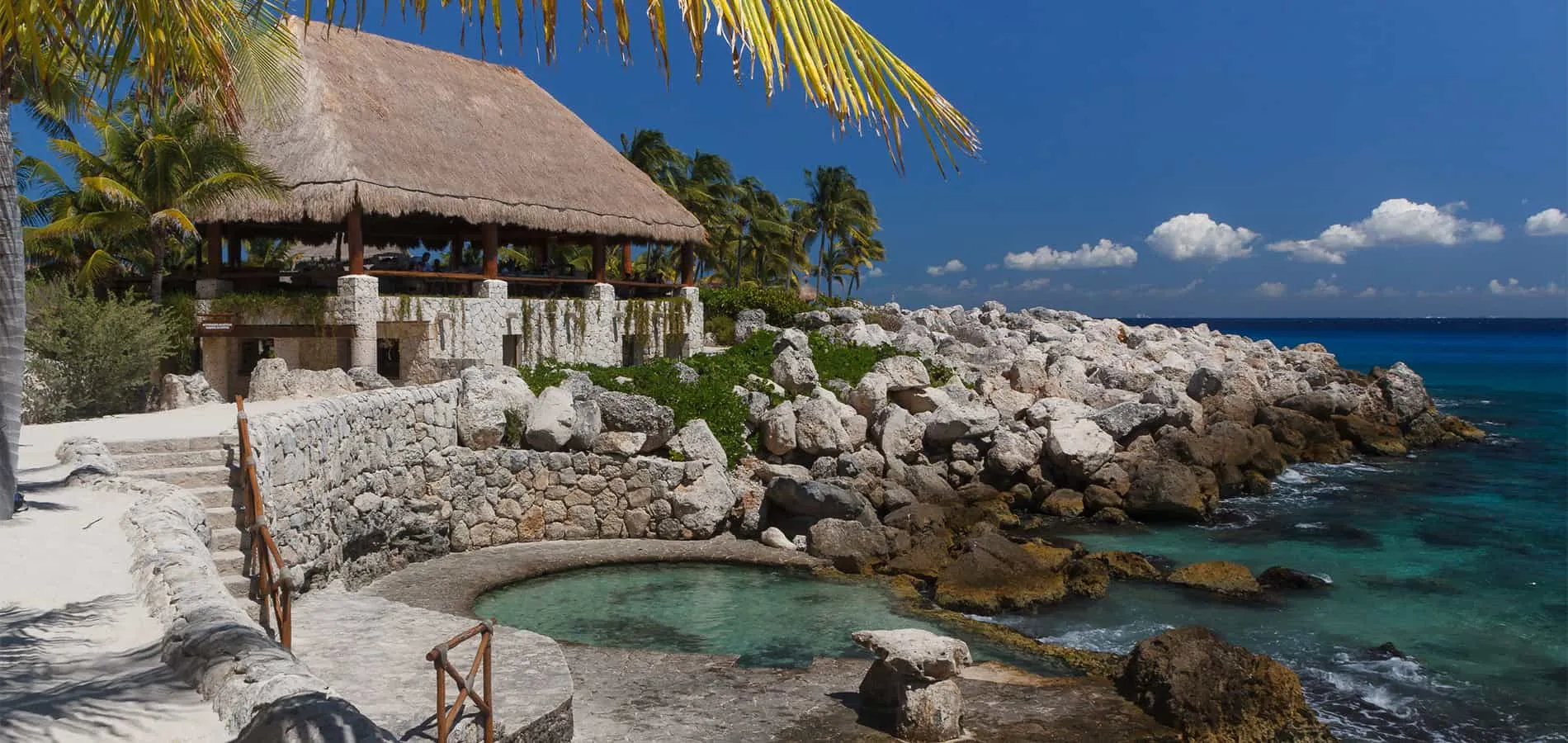 Palapa junto al mar con rocas, piscina natural y palmeras al fondo.