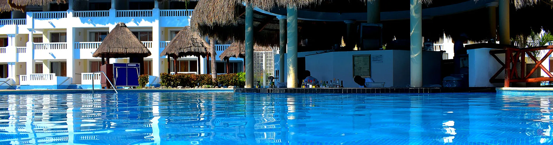 Piscina con bar y sombrillas de paja en un hotel, con edificios al fondo.