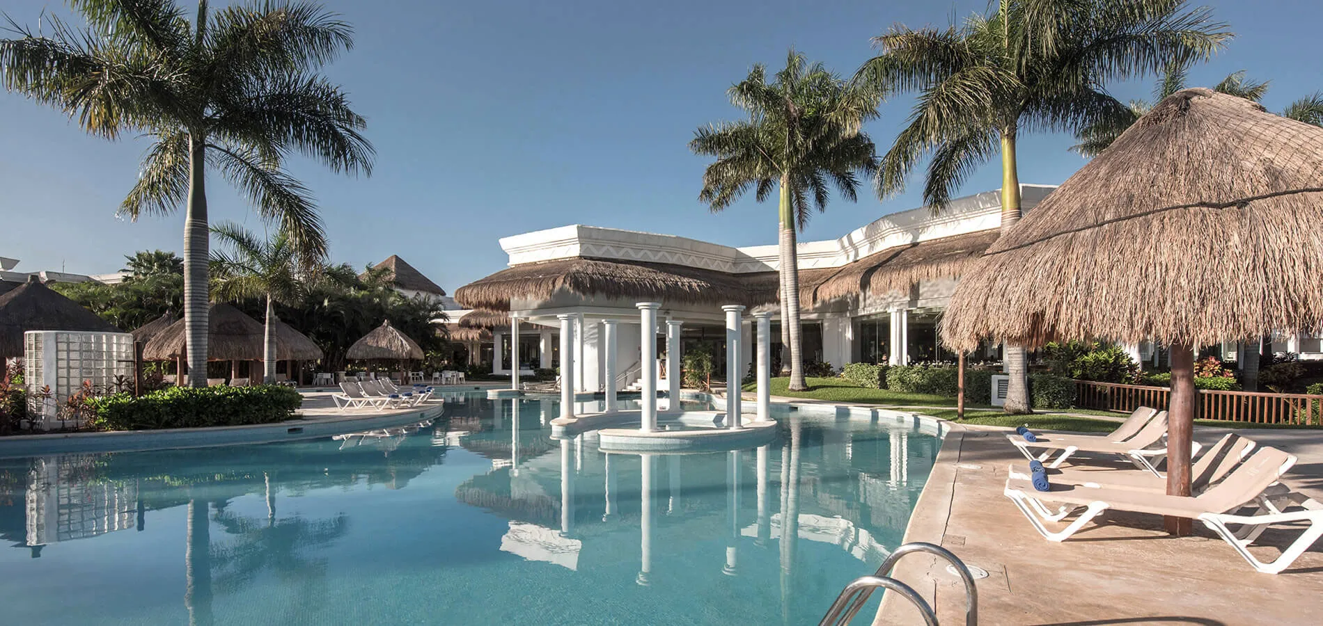 Piscina con palmeras y tumbonas junto a un edificio con techo de palapa y columnas blancas.