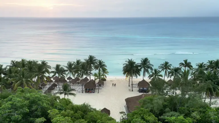 Una amplia vista de una playa tropical con arena blanca, palmeras, cabañas con techo de paja y aguas tranquilas de color turquesa bajo un cielo nublado.