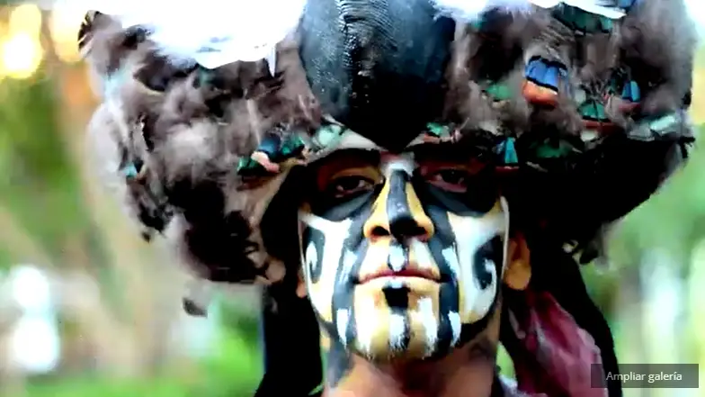 Persona con pintura facial tribal y elaborado tocado con plumas, en fondo desenfocado.
