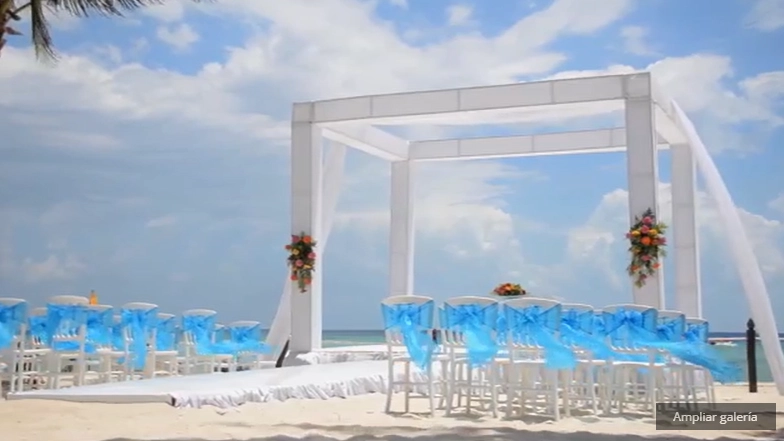 Boda en la playa con sillas blancas adornadas con lazos azules y un altar bajo un cielo azul.