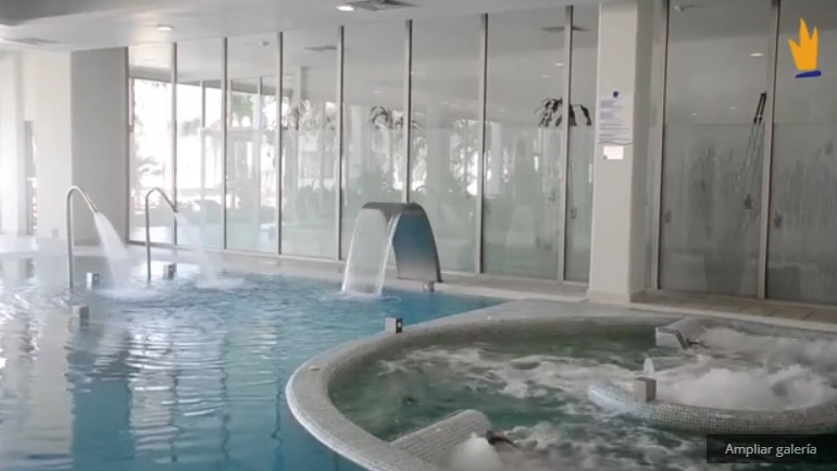 Piscina interior con chorros de agua y jacuzzi, rodeada de paredes acristaladas.