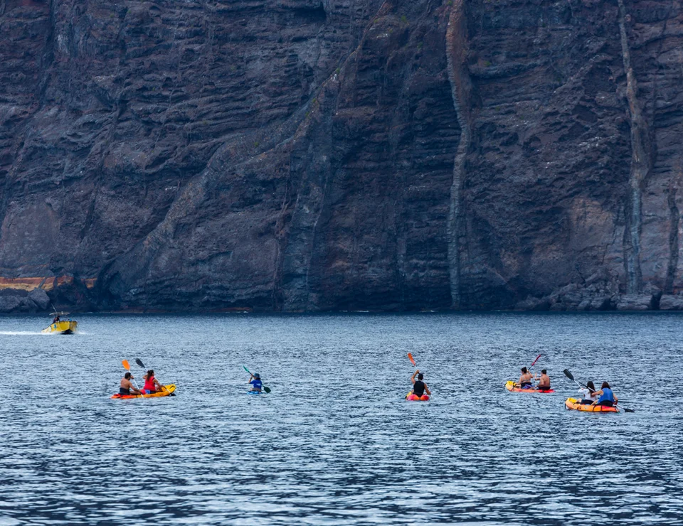Personas en kayaks y un bote amarillo cerca de un imponente acantilado rocoso.