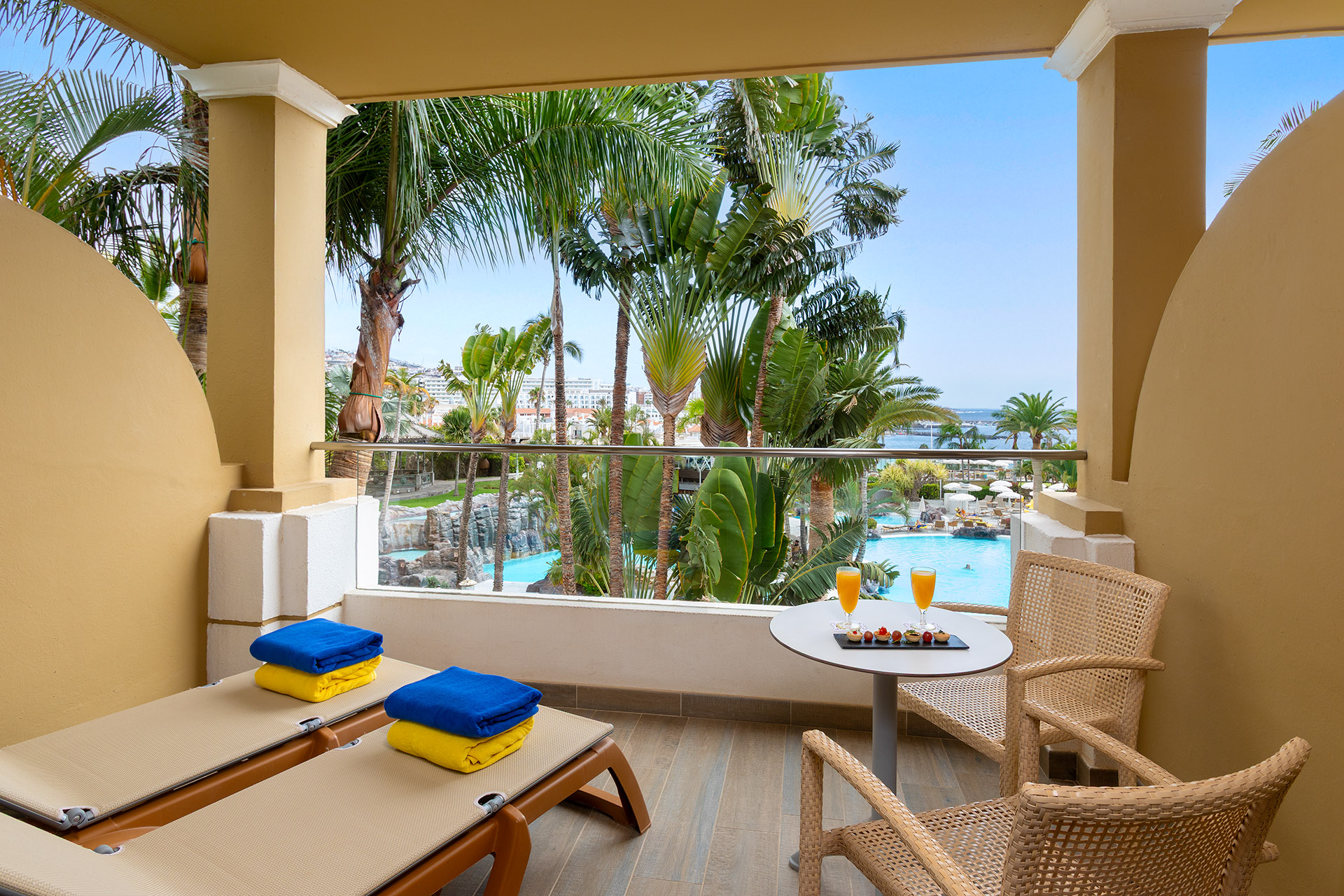 Terraza con vista a piscina y jardines, dos sillas, mesa con jugo y toallas.