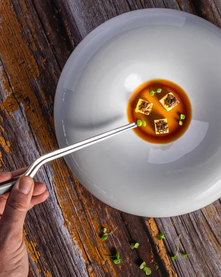 Plato de sopa con cubos y hierbas, servido con una pipeta sobre mesa de madera.