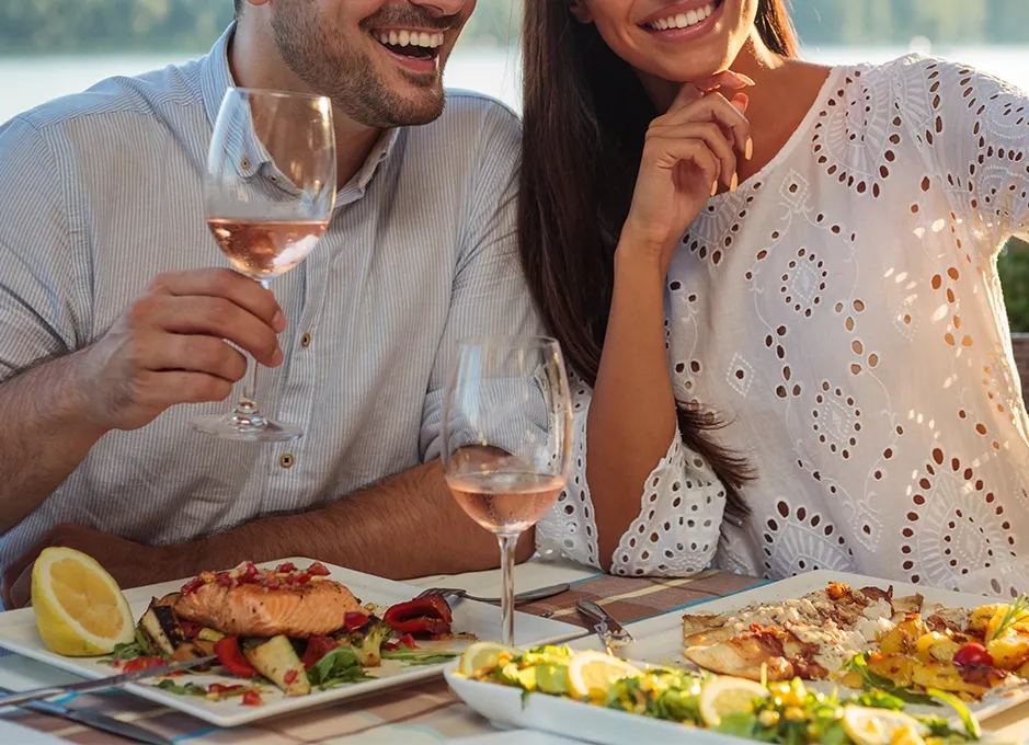 Pareja sonríe brindando con vino rosado en una cena al aire libre, platos de pescado y verduras.