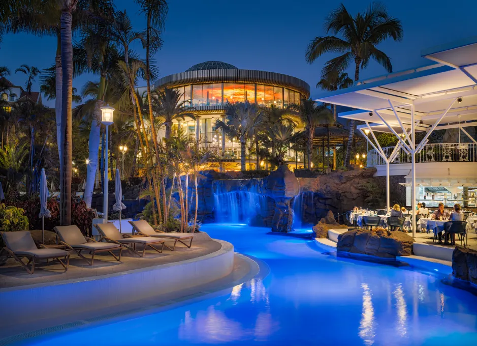 Piscina iluminada de noche con cascada, rodeada de palmeras y restaurante.