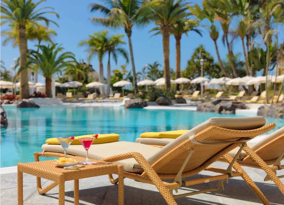 Piscina rodeada de palmeras con tumbonas y bebidas en primer plano, bajo un cielo despejado.