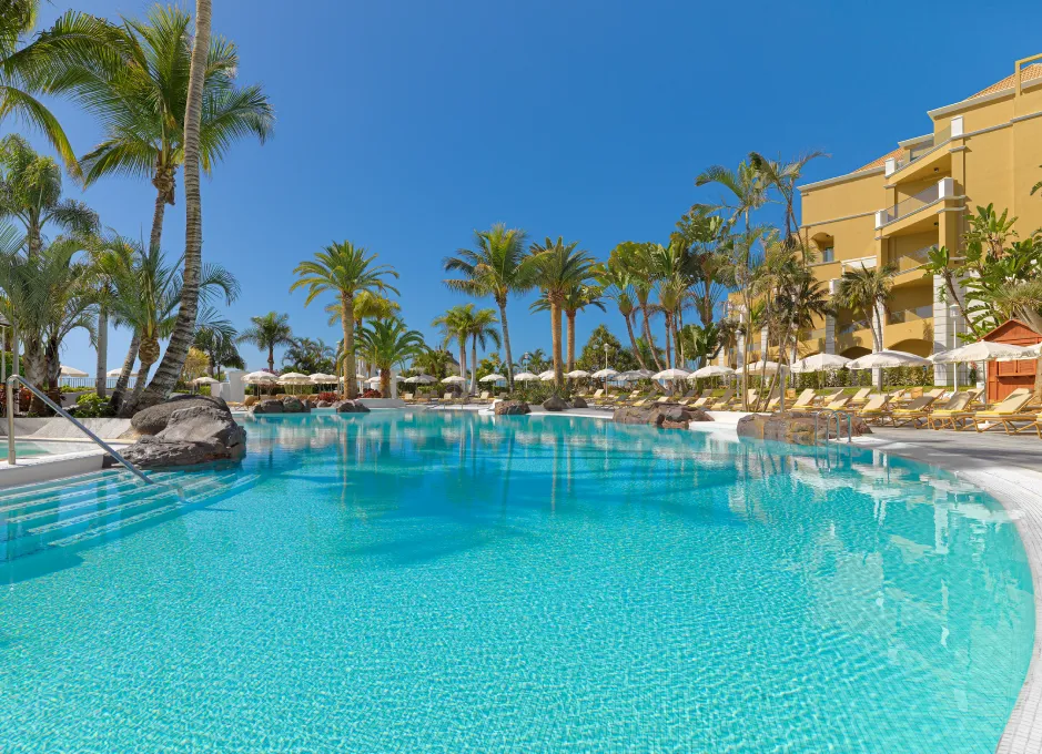 Piscina de hotel rodeada de palmeras y tumbonas bajo un cielo despejado y azul.