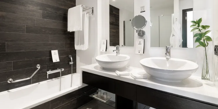 Baño moderno con dos lavabos, espejo, bañera, toallas blancas y planta decorativa.