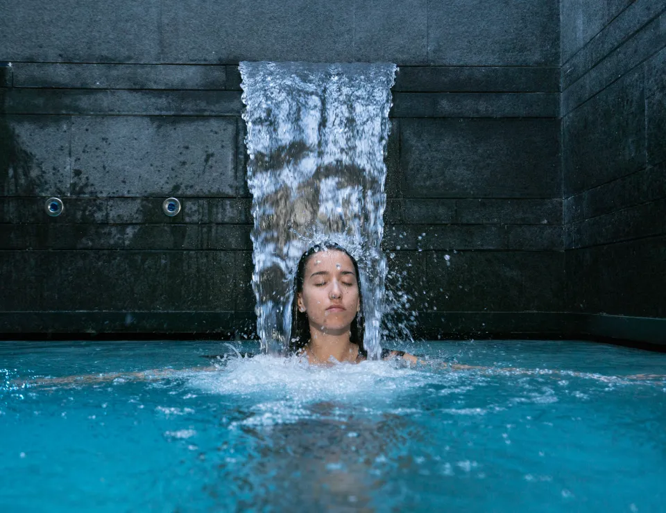 Persona bajo una cascada en una piscina, con los ojos cerrados y expresión relajada.