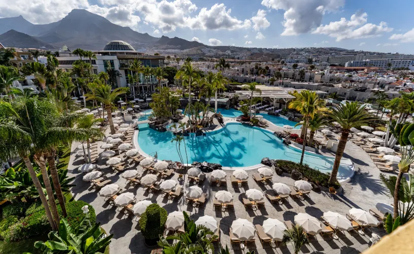 Vista de una piscina en un resort tropical, rodeada de palmeras y montañas al fondo.