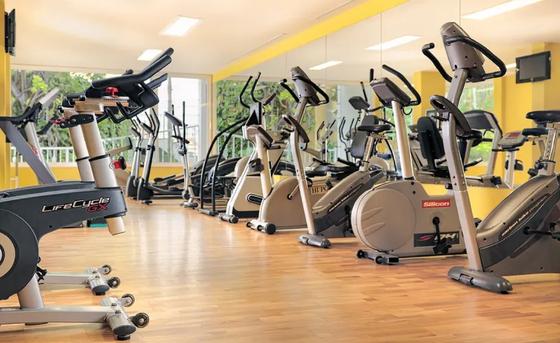 Gimnasio con bicicletas estáticas frente a un espejo, suelo de madera y paredes amarillas.