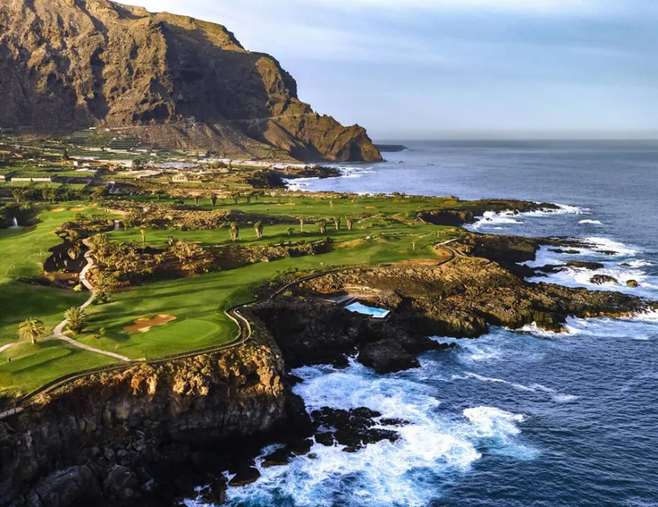 Costa rocosa con campos verdes y montañas al fondo, junto a un océano con olas espumosas.