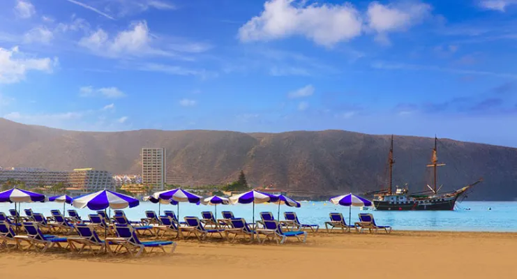 Playa con sombrillas y tumbonas frente al mar, barco al fondo y colinas bajo un cielo azul.