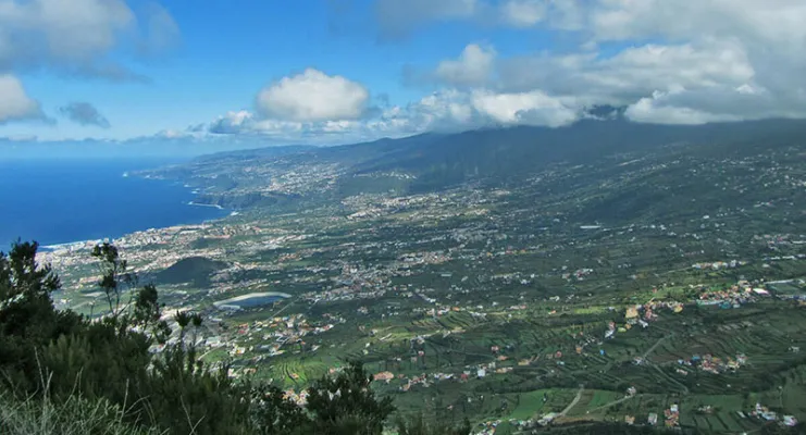 Vista panorámica de un paisaje costero con montañas, vegetación y un cielo parcialmente nublado.