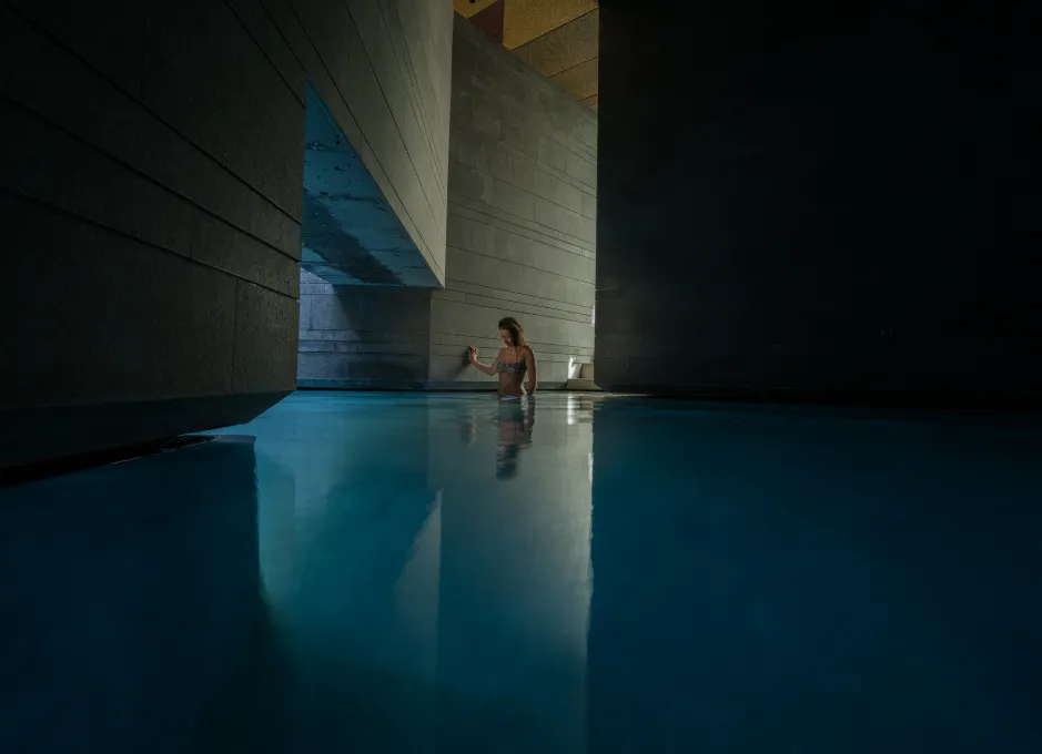 Persona en una piscina interior iluminada, rodeada de paredes de cemento y luz tenue.
