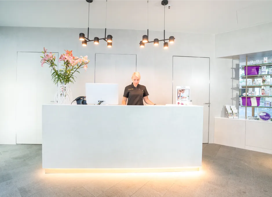 Recepción moderna con una persona detrás del mostrador y flores a la izquierda.