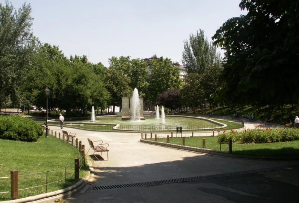 Fuente rodeada de césped y árboles en un parque, con bancos y personas caminando.