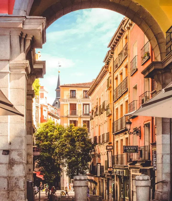 Calle colorida con edificios antiguos y árbol, vista a través de un arco de piedra bajo un cielo despejado.