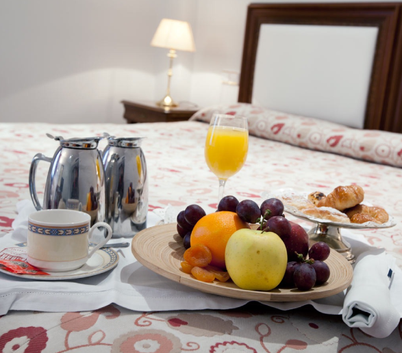 Desayuno en cama: café, jarras de metal, jugo de naranja, frutas variadas y bollos sobre una cama decorada.
