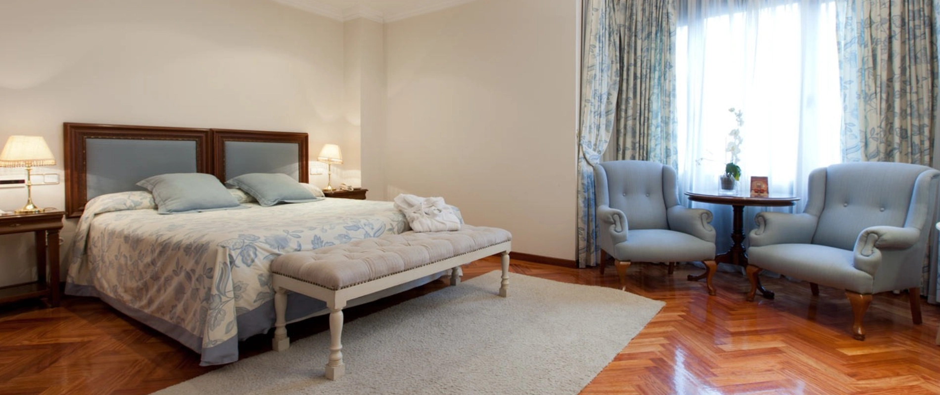 Dormitorio elegante con cama doble, sillones azules, mesita, lámparas y cortinas. Piso de madera y decoración clásica.