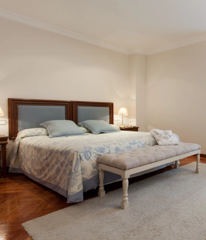 Habitación con cama grande, ropa de cama floral, cojines azules, lámparas de noche y un banco acolchado enfrente.