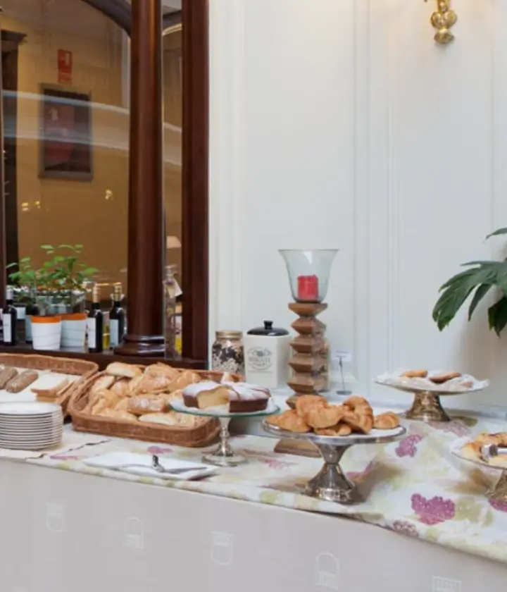 Mesa con variedad de panes y pasteles sobre mantelería floral en un entorno elegante con plantas y decoración cálida.