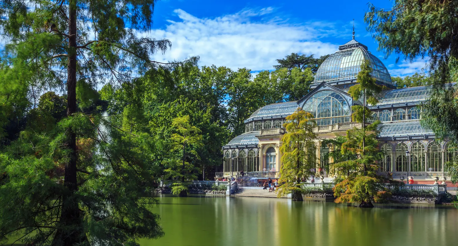 Edificio de cristal con cúpula rodeado de árboles y un lago, bajo un cielo azul.