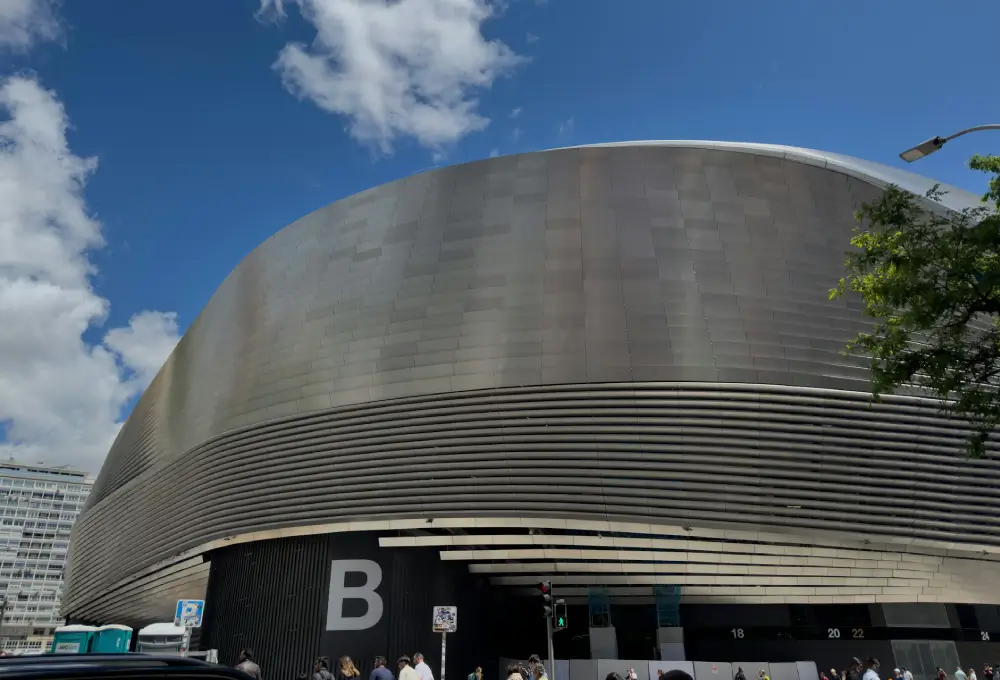 Edificio moderno con fachada curva y metálica, letra "B" en la entrada, cielo azul con algunas nubes.