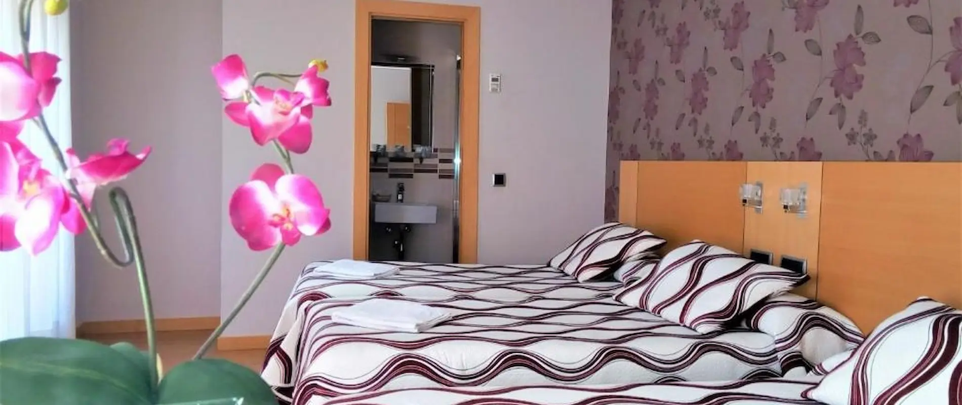 Habitación con dos camas, colcha a rayas, orquídeas rosas y fondo floral en la pared.