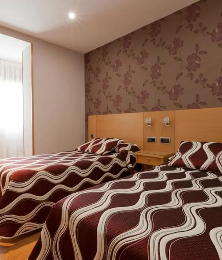Dos camas con colchas de ondas marrones en una habitación con papel tapiz floral y luz natural.