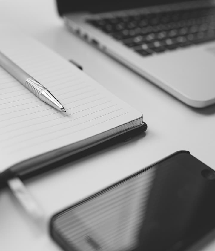 Cuaderno con bolígrafo, junto a una laptop y un celular sobre un escritorio. Blanco y negro.