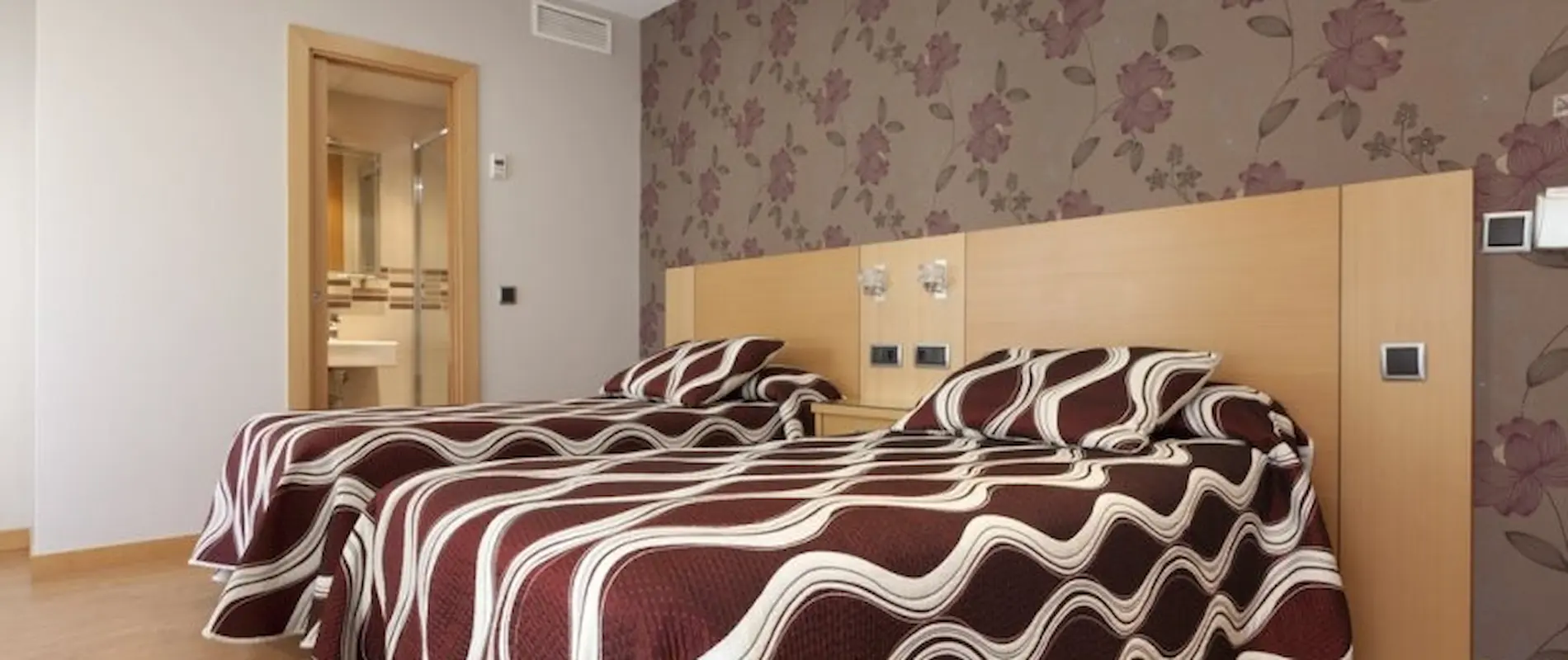 Dos camas con colchas geométricas en habitación con pared floral y baño al fondo.