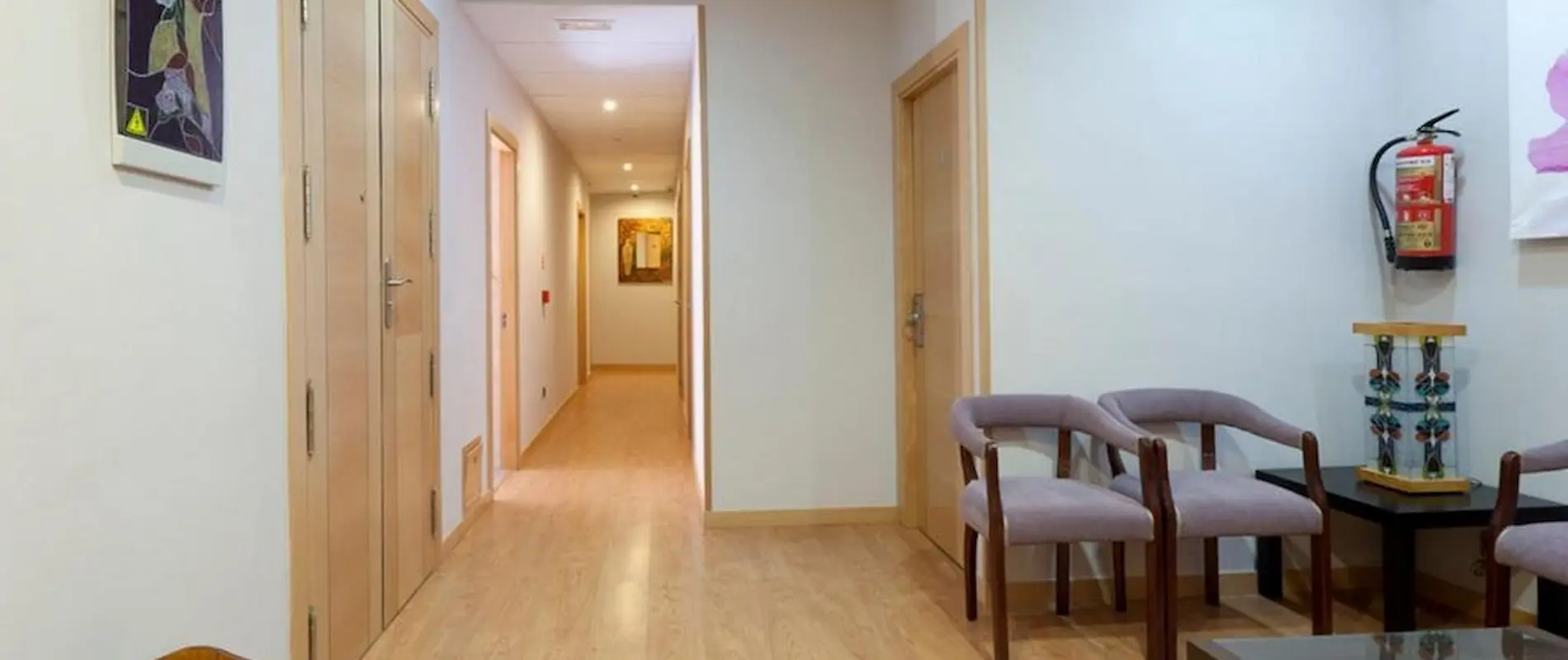 Pasillo con suelo de madera, puertas, sillas y extintor en la pared.