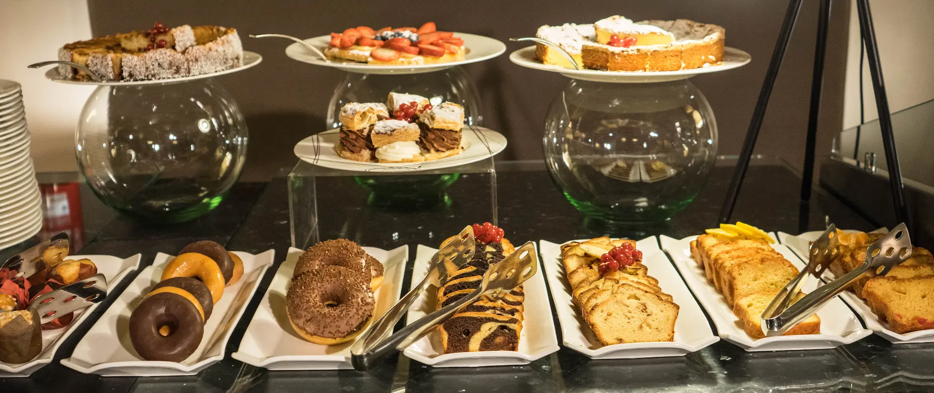 Variedad de postres en un buffet: tartas, donuts y bizcochos en platos blancos.