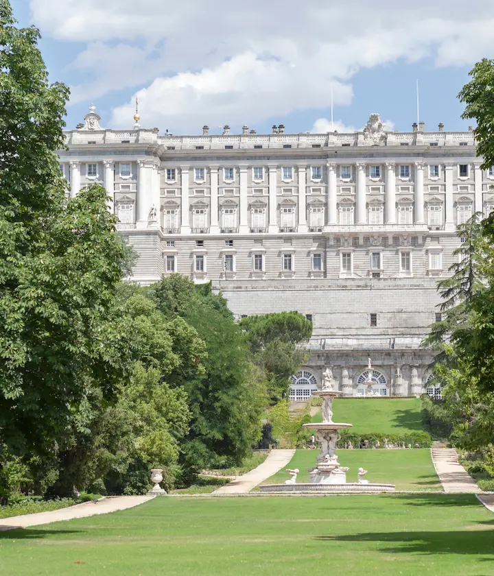 Palacio Real de Madrid visto desde un jardín con árboles y una fuente central.