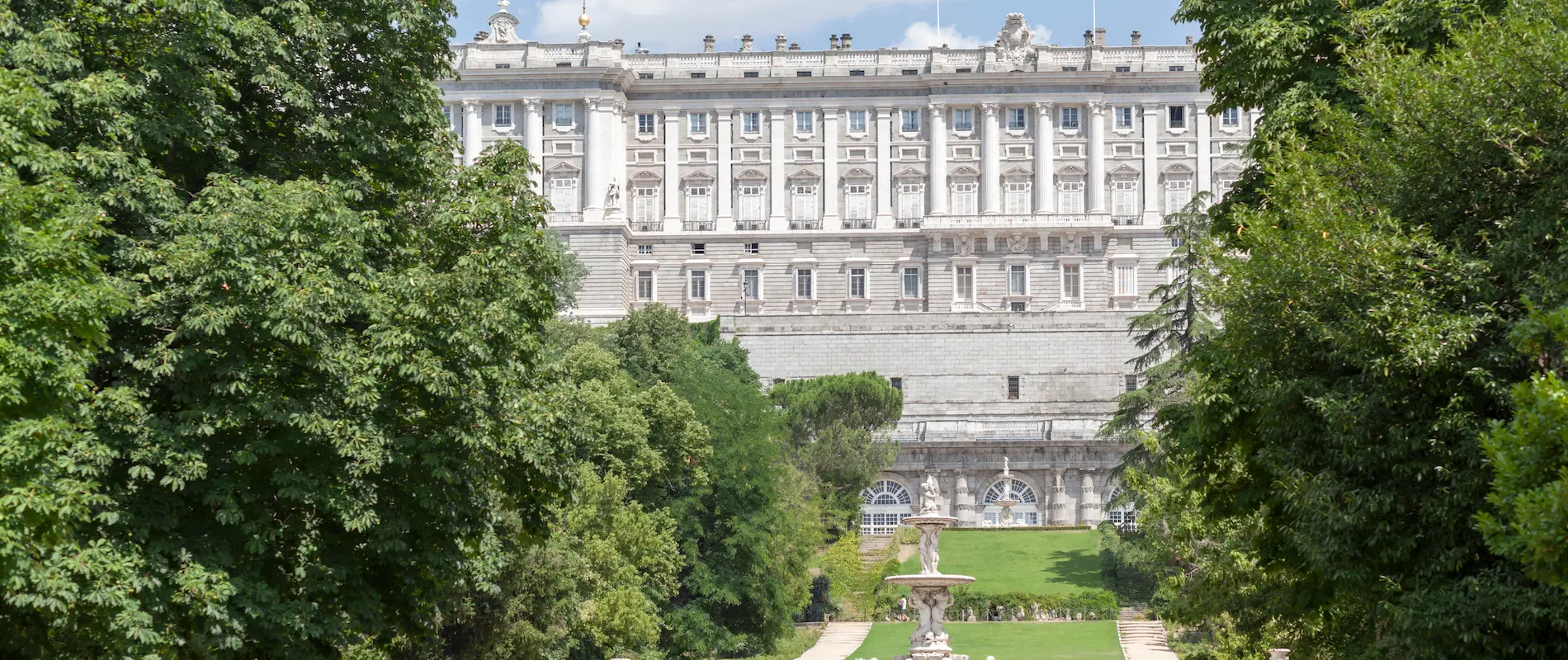 Palacio entre árboles, con fuente en jardín frontal y cielo azul de fondo.